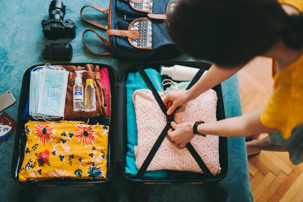 An overhead view of a young woman packing her suitcase for vacation, including a stack of mask along with her passport, clothes and camera.