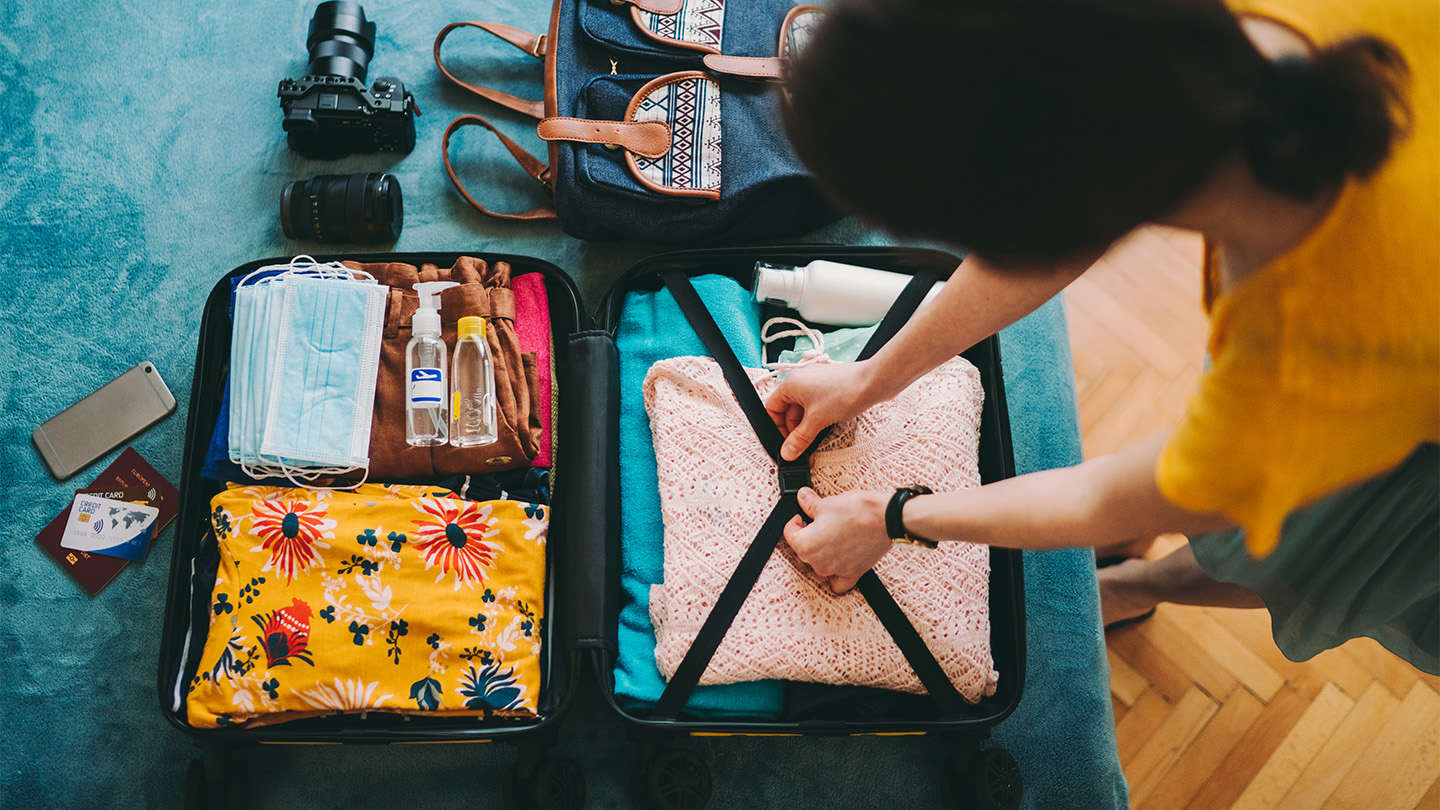 An overhead view of a young woman packing her suitcase for vacation, including a stack of mask along with her passport, clothes and camera.