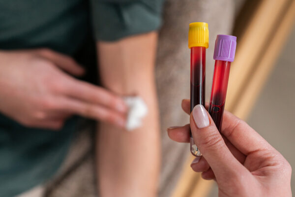 A stock photograph of a nurse holding two tubes of blood. In the background, a patient in a green shirt holds a cotton ball to their arm after having blood drawn.