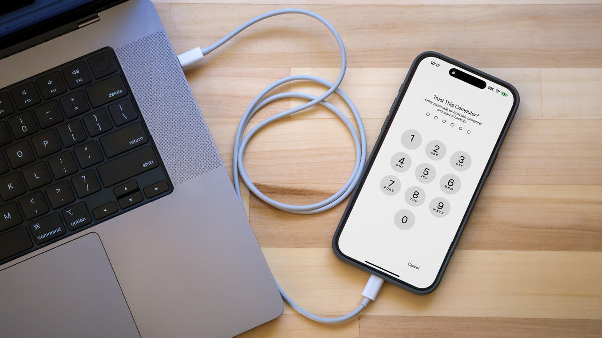 Top-down view of an iPhone connected to a MacBook Pro via a white cable. The iPhone&apos;s screen shows the number pad for entering a code.