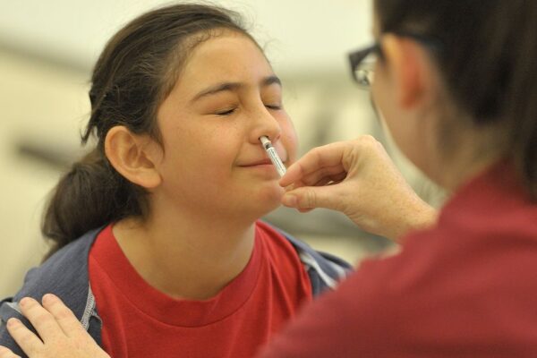 Girl has her eyes shut as a healthcare provider sprays a nasal vaccine up her right nostril