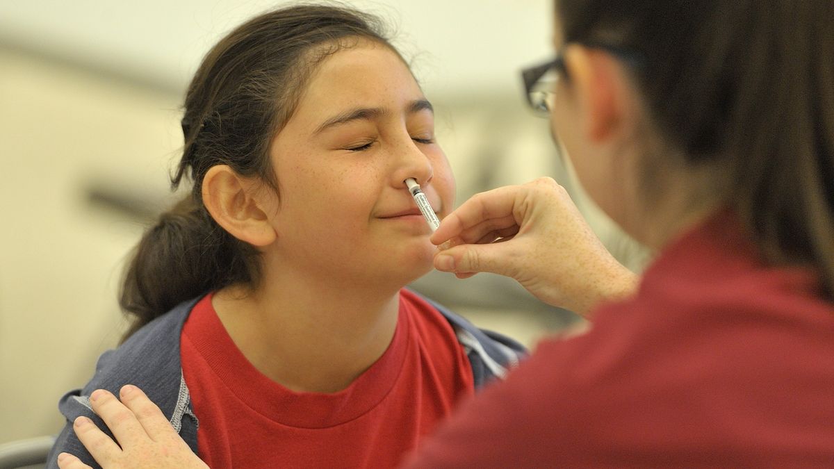 Girl has her eyes shut as a healthcare provider sprays a nasal vaccine up her right nostril