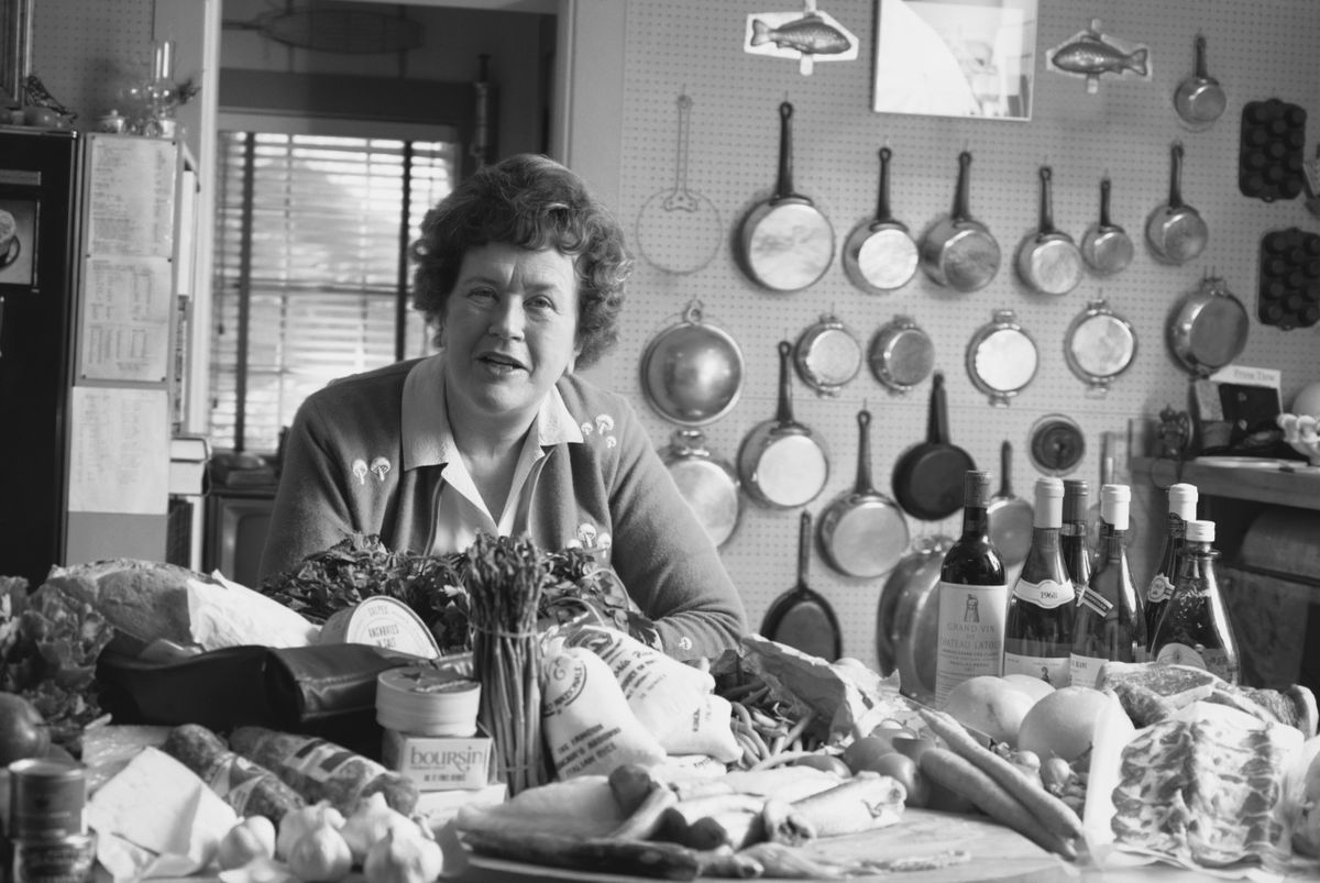 A black and white photo of Julia Childs with an array of fresh ingredients in the kitchen