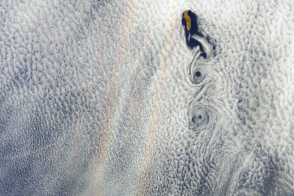 A satellite photo showing a faint double band of rainbow next to an island with cloud swirls forming off its coastline