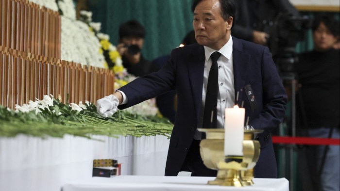 A man in formal attire, wearing a white glove, places flowers at a memorial altar adorned with white chrysanthemums. Candles are lit in the foreground, and a backdrop of flowers and wooden memorial tablets is visible.